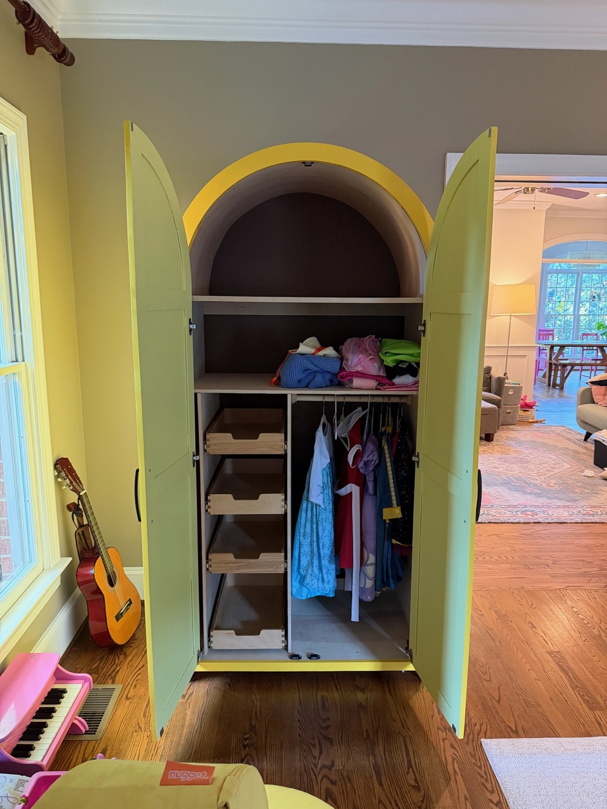Open armoire showing four wooden drawers on the left, dress-up clothes hanging on a rod on the right, and shelves above