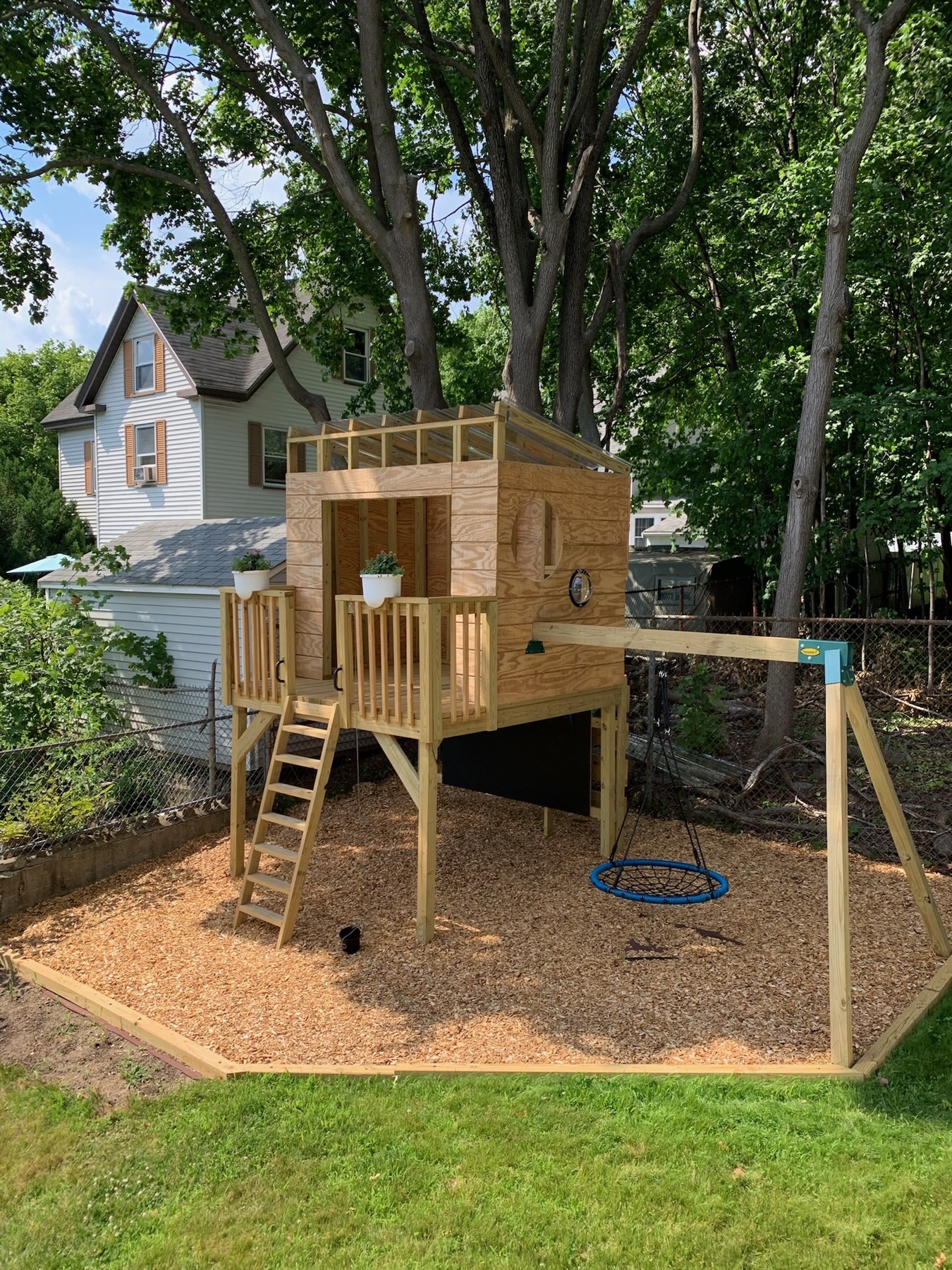 Custom wooden backyard play structure with raised playhouse, ladder, porch railings and blue nest swing